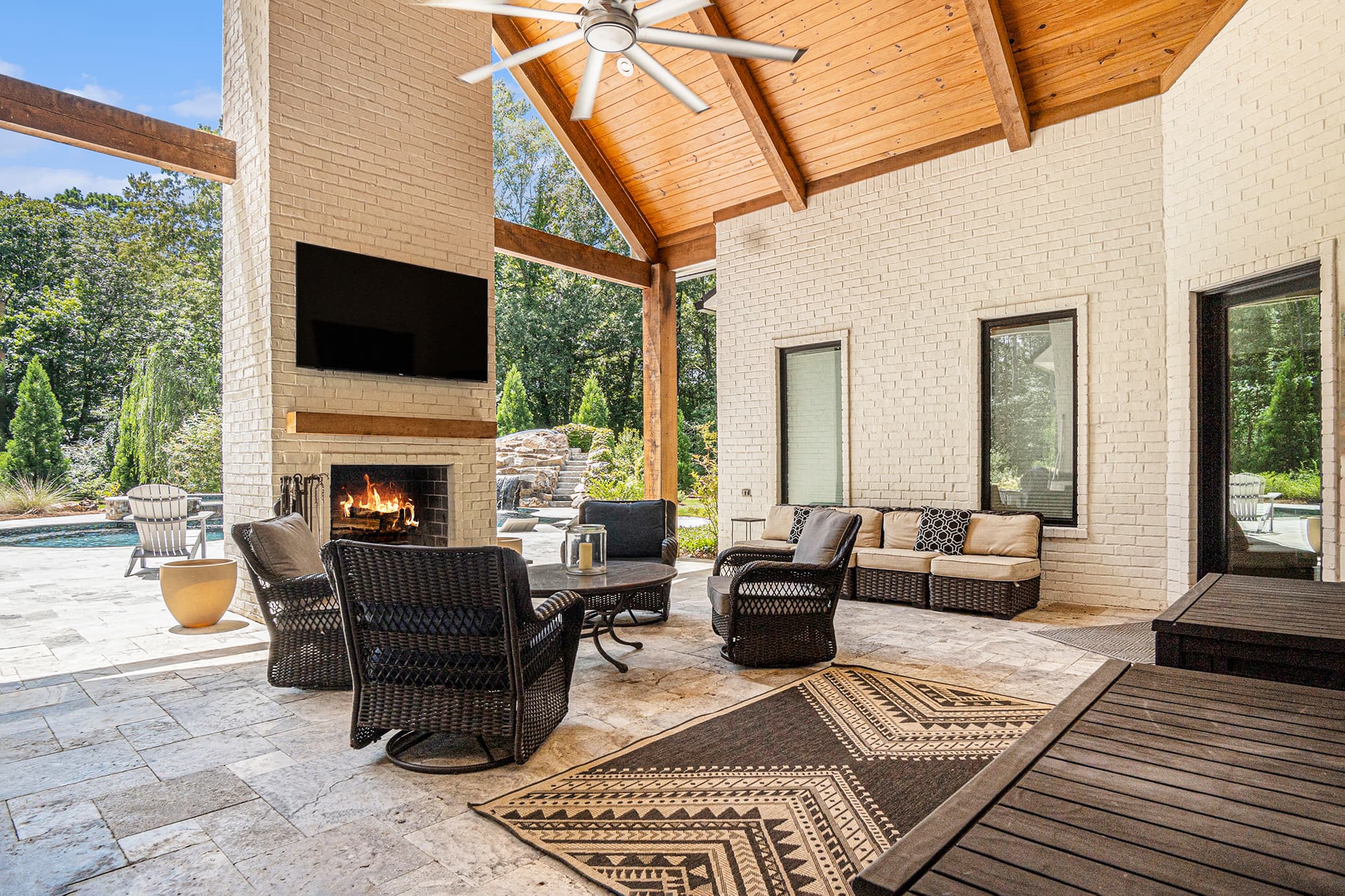Covered outdoor living room with a brick fireplace, vaulted wood ceiling, and pool beyond